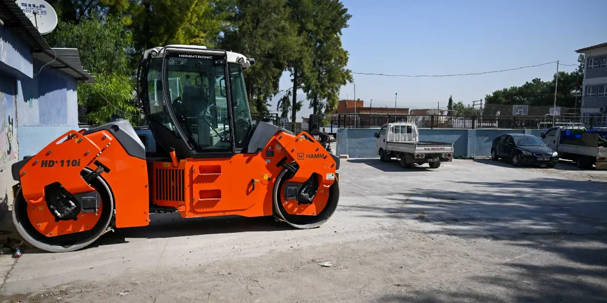 Açık havada, beton bir zemin üzerinde park halinde duran turuncu renkli, Hamm marka büyük bir silindir iş makinesi. İş makinesinin arka planında beyaz bir kamyonet, siyah bir binek otomobil ve başka bir kamyonet daha bulunuyor. Çevrede ağaçlar, bir duvar ve binalar yer alıyor.