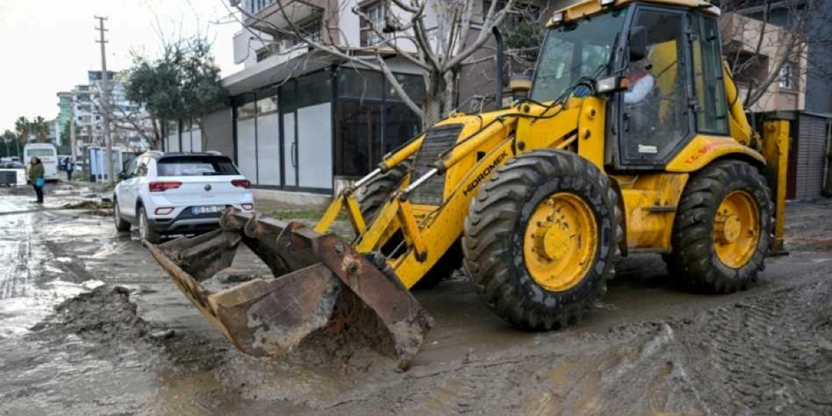 Bornova'da sel sonrası çamurla kaplanmış bir sokakta, üzerinde Hidromek yazan sarı renkli bir iş makinesi çalışma yapıyor. İş makinesinin hemen yanında beyaz bir otomobil park halinde duruyor, arka planda ise binalar ve yürüyen insanlar görülüyor.