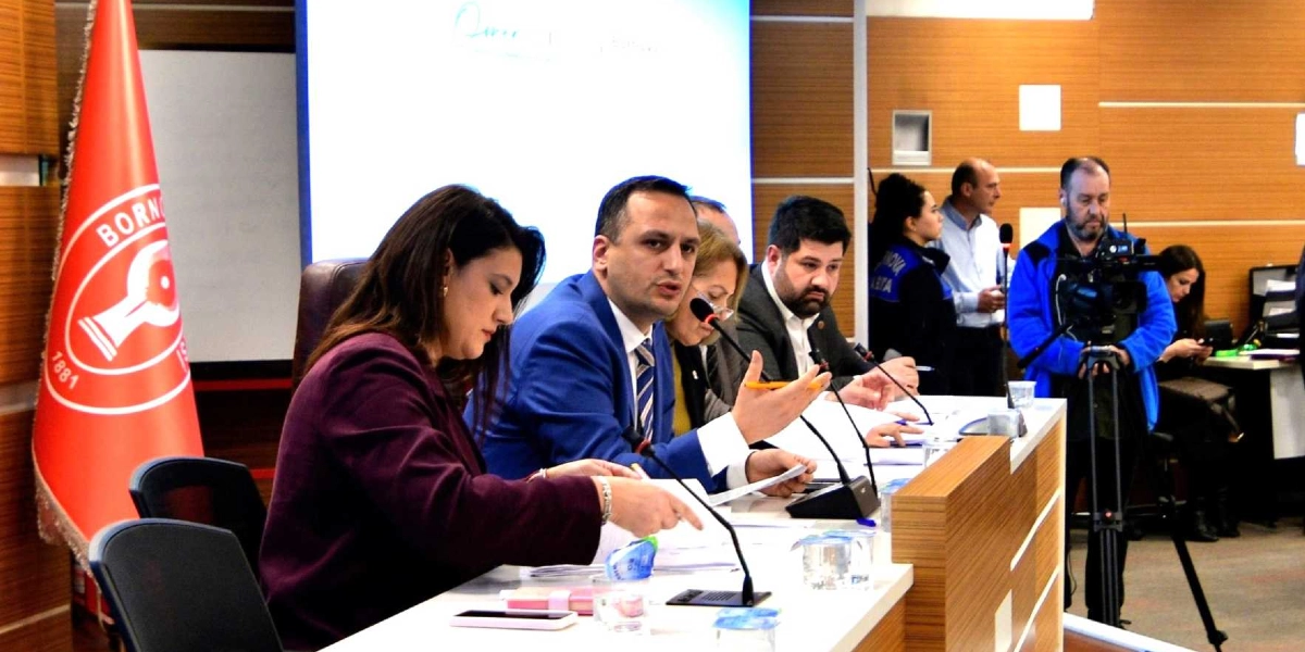 Bornova Belediyesi'ne ait bir toplantı salonunda, masada oturan yetkililer bir sunum veya toplantı gerçekleştiriyor. Ön planda, koyu renkli bir ceket giyen kadın ve yanında mavi ceketli, kravatlı bir erkek konuşma yapıyor. Masanın üzerinde mikrofonlar, belgeler ve su bardakları bulunuyor. Arka planda Bornova Belediyesi'nin kırmızı renkli bayrağı yer alırken, sağ tarafta bir kameraman çekim yapıyor.