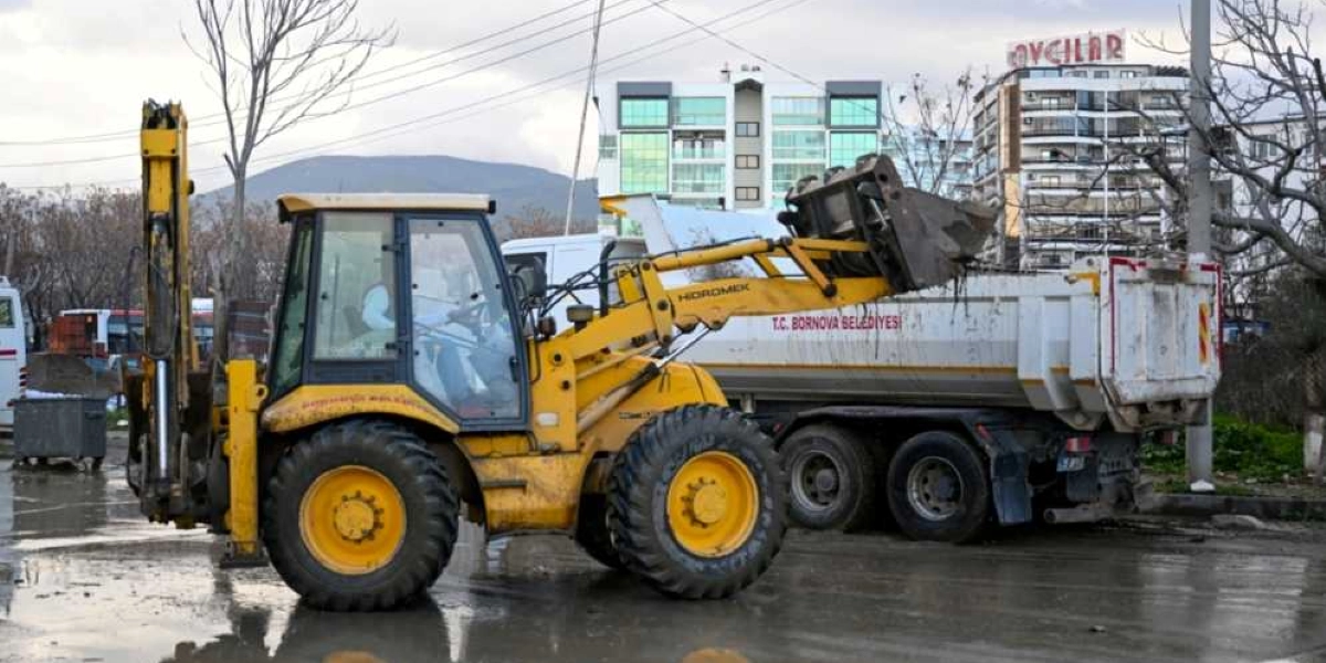 T.C. Bornova Belediyesi'ne ait sarı renkli bir iş makinesi, ıslak ve çamurlu bir zeminde, üzerinde yine belediyeye ait yazıların bulunduğu bir kamyonun kasasına toprak veya moloz boşaltıyor. Arka planda çok katlı binalar ve ağaçlar görülüyor.