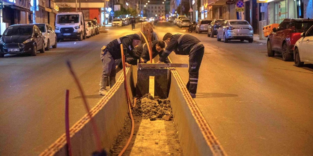 Gece vakti aydınlatılmış bir şehir caddesinde, yolun ortasında açılmış uzun bir kanal üzerinde çalışan üç işçi. İşçiler, kanalın içine yerleştirilen altyapı boruları ve kablolar üzerinde düzenleme yapıyor. Çevrede park halindeki araçlar ve boş bir cadde görülüyor.