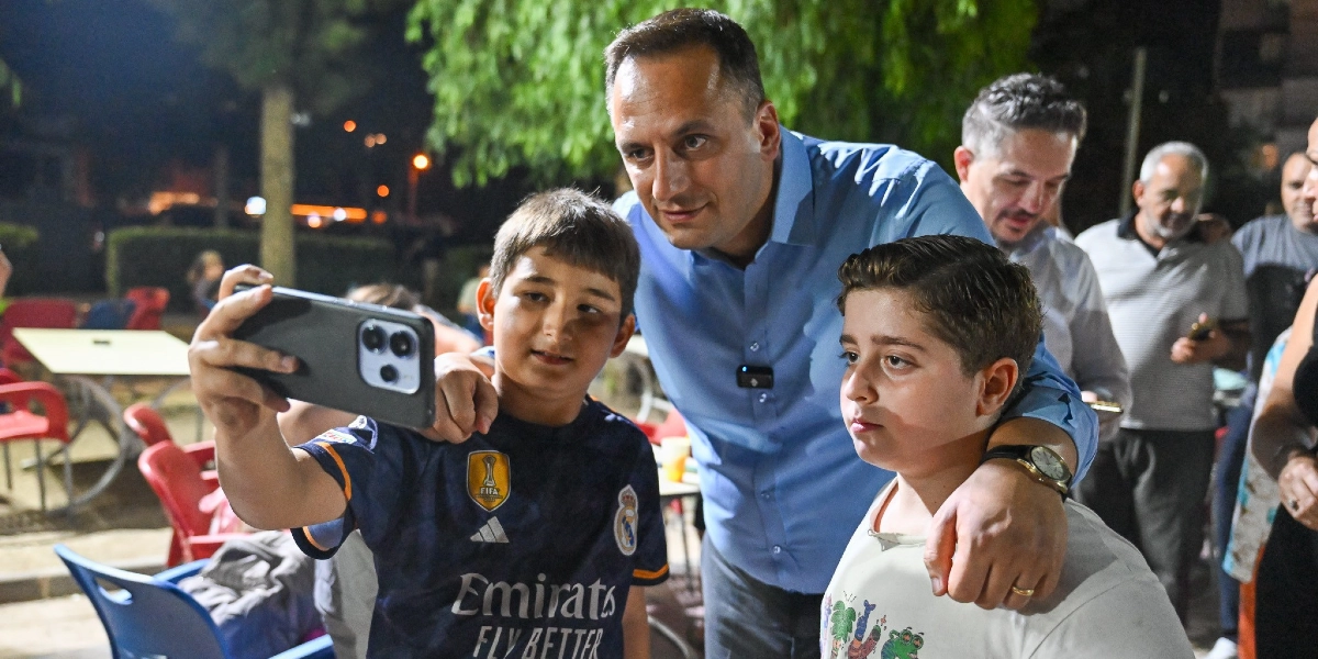 Mavi gömlekli bir erkek, akşam saatlerinde dış mekanda iki çocukla birlikte selfie çekiliyor. Çocuklardan biri Real Madrid forması giyiyor ve telefonu tutuyor, diğeri ise yanlarında duruyor. Başkan, çocukların omuzlarına elini atarak onlarla birlikte kameraya gülümsüyor. Arka planda ise masa ve sandalyelerin bulunduğu bir park alanı görülüyor.