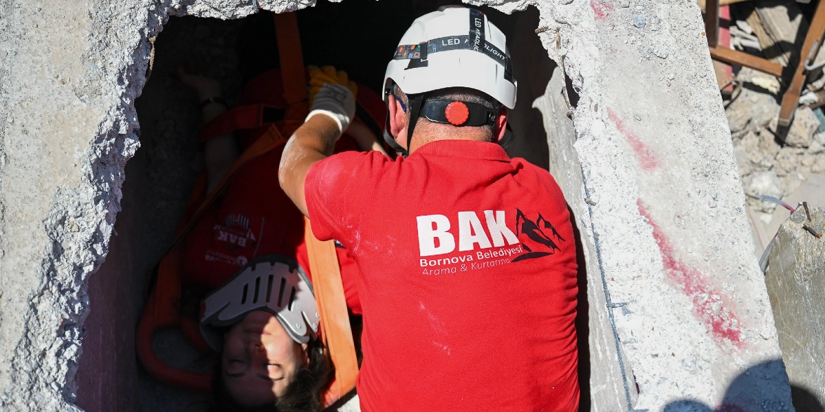 Bornova Belediyesi Arama ve Kurtarma (BAK) ekibinden beyaz baretli bir kurtarma görevlisi, enkaz altındaki dar bir boşlukta boyunluk takılı olan bir depremzedeyi sedye yardımıyla dışarı çıkarmaya çalışıyor.