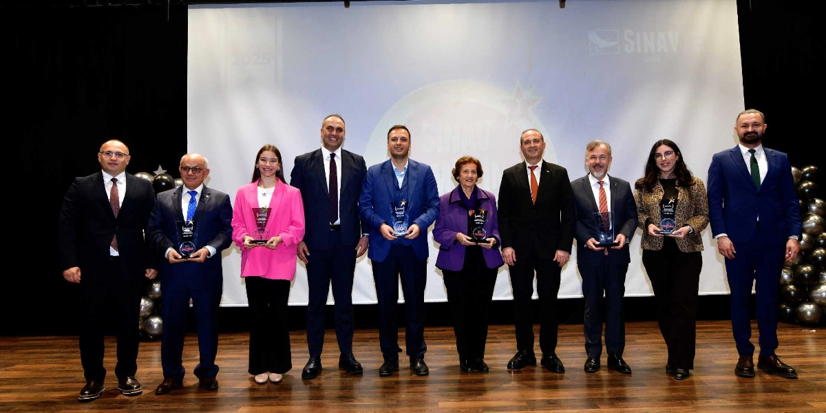 Bir sahnede yan yana dizilmiş, ellerinde plaketler tutan dokuz kişilik bir grup. Arka planda üzerinde 