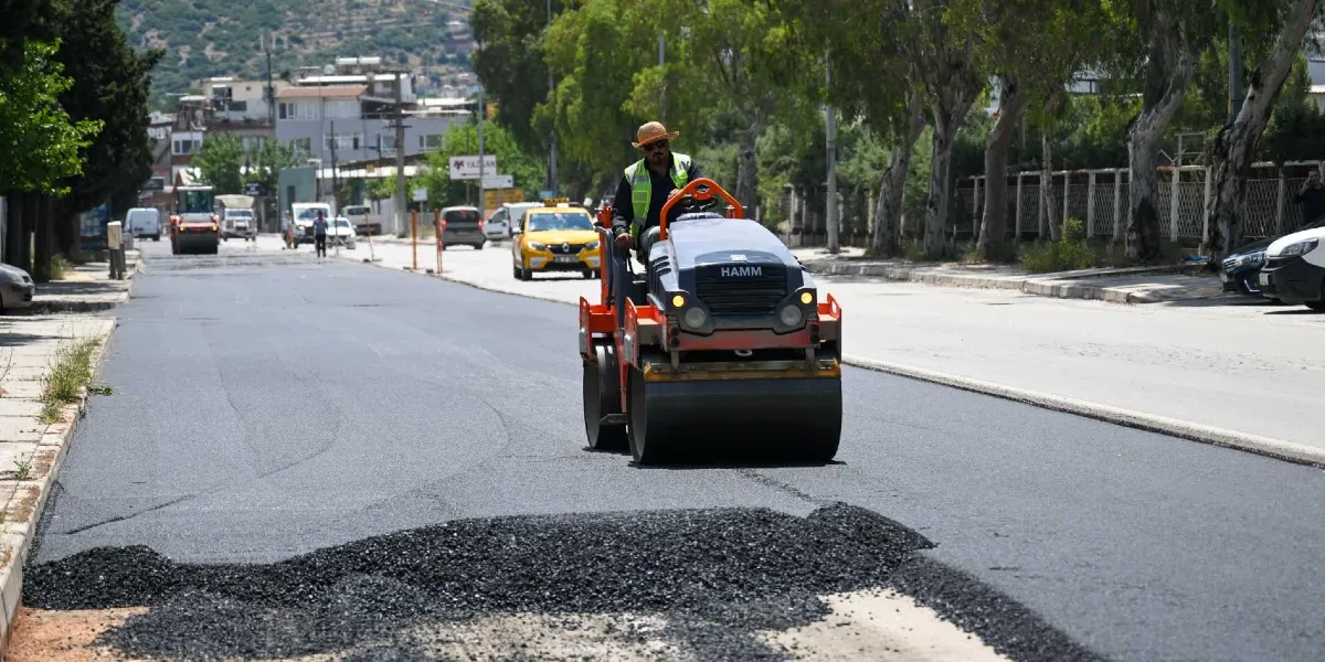 Pınar Caddesi'nde devam eden yol yenileme çalışmaları kapsamında, üzerinde Hamm marka bir silindir operatörünün bulunduğu iş makinesi, yeni dökülmüş asfaltı düzleştiriyor. Arka planda ise trafik akışının devam ettiği caddede park halindeki araçlar ve ağaçlıklı bir yol kenarı görülüyor.