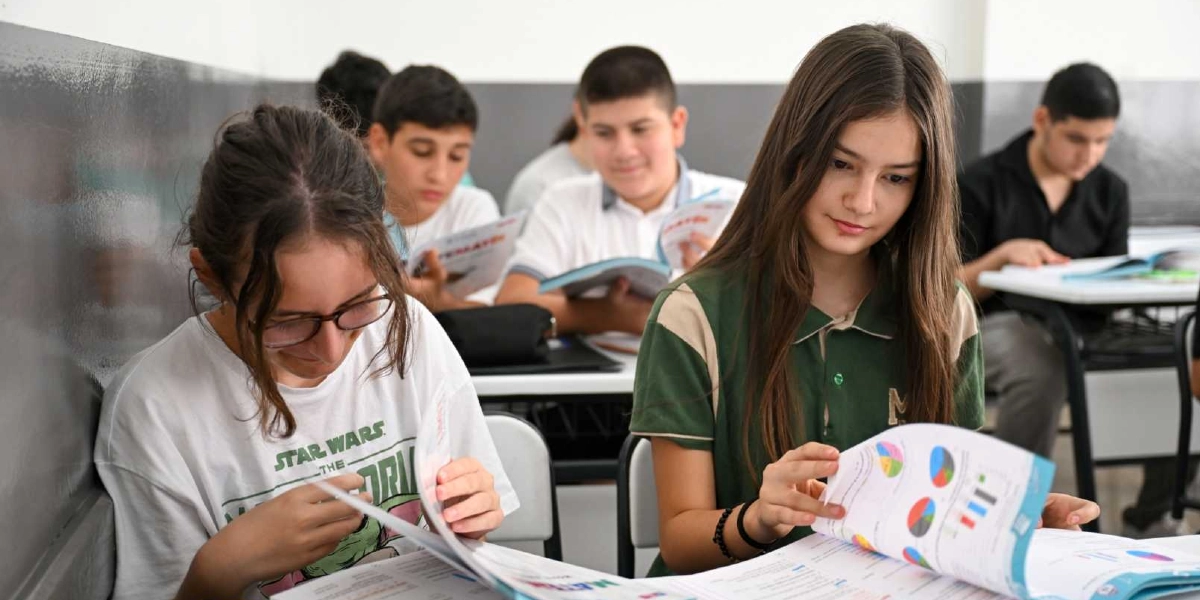 Sınıf ortamında sıralarında oturan ve önlerindeki ders kitaplarını inceleyen ortaokul veya lise çağındaki öğrenciler. Ön planda, beyaz tişörtlü gözlüklü bir kız öğrenci ile yeşil yakalı bir forma giyen uzun saçlı bir kız öğrenci dikkatle kitaplarını okuyor; arka planda ise diğer öğrenciler ders çalışmaya devam ediyor.