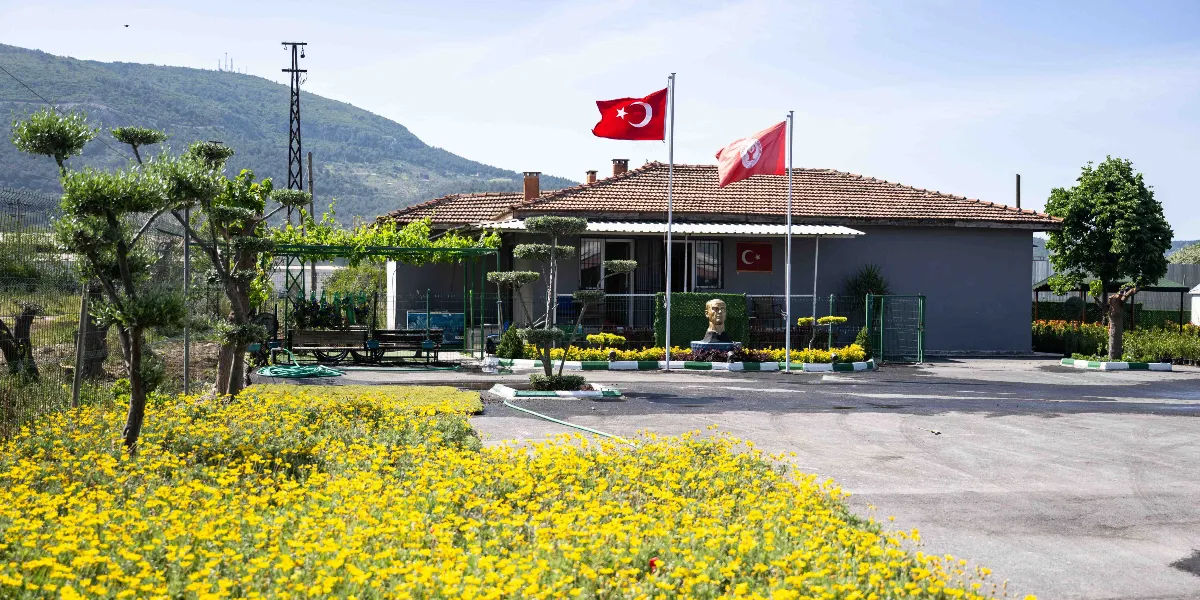 Arka planda dağ manzarası olan, önünde sarı çiçeklerle dolu geniş bir bahçenin bulunduğu tek katlı, kiremit çatılı bir yapı. Binanın önündeki direklerde Türk bayrağı ve üzerinde ay yıldız bulunan kırmızı bir flama dalgalanıyor. Bahçede Atatürk büstü, düzenli budanmış ağaçlar ve yeşil bir çardak yer alıyor.