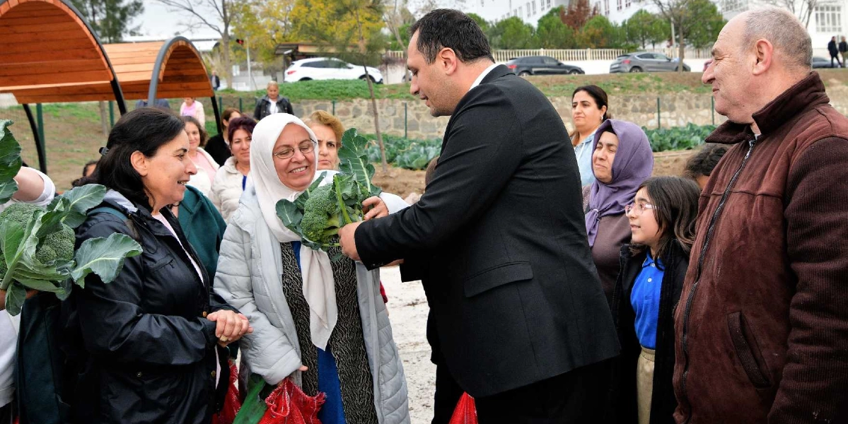Bornova'daki bir kent bostanında, siyah takım elbiseli bir erkek, hasat edilen brokolileri beyaz başörtülü ve açık renkli montlu bir kadına uzatıyor. Çevredeki diğer vatandaşlar ve bir çocuk, gülümseyerek bu anı izliyor. Arka planda ise ahşap çatılı oturma alanları ve tarım yapılan bostan alanı görülüyor.