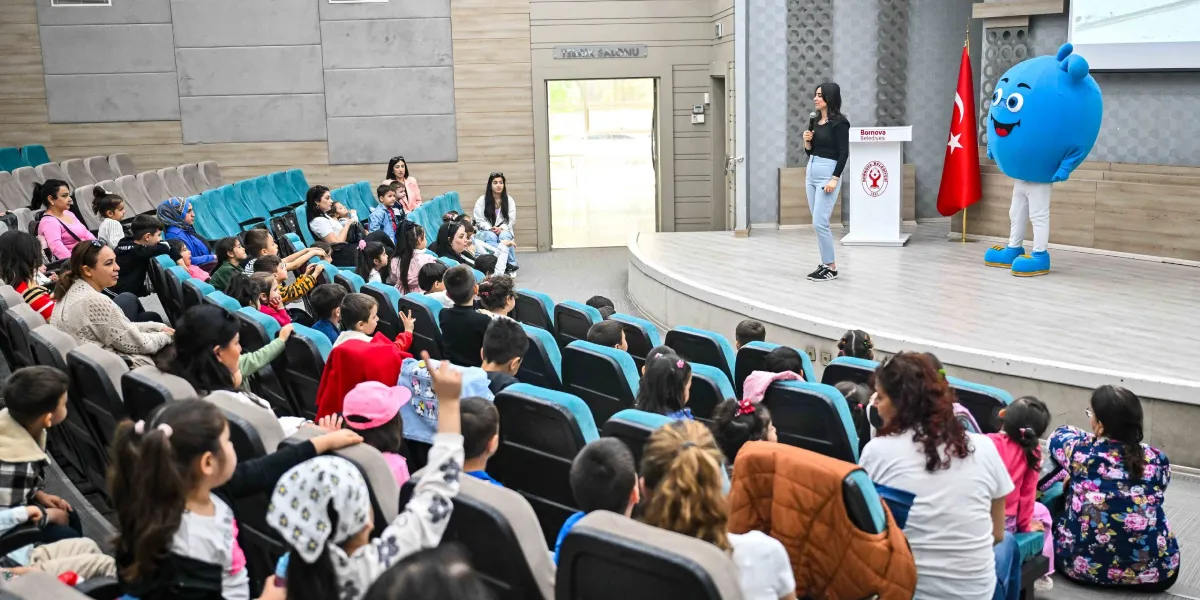 Modern bir konferans salonunda, sahnede mikrofonla konuşan bir kadın ve yanında Bornova Belediyesi logolu kürsünün önünde duran büyük, mavi, maskot bir karakter bulunuyor. Salonun koltuklarında oturan çok sayıda çocuk ve onlara eşlik eden yetişkinler, sahnedeki sunumu ilgiyle izliyor. Sahnenin sağ tarafında bir Türk bayrağı yer alıyor.