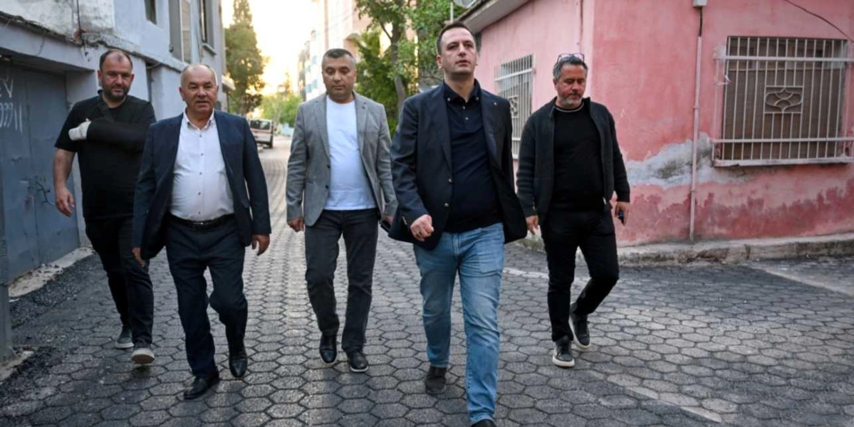 Beş kişilik bir erkek grubu, Egemenlik Mahallesi'ndeki parke taşlı bir sokakta yan yana yürüyerek ilerliyor. Grubun önünde yer alan Başkan Eşki, lacivert blazer ceket, koyu renkli tişört ve kot pantolon giyiyor. Yanındaki diğer kişiler ise günlük kıyafetler ve ceketlerle onlara eşlik ediyor. Arka planda sol tarafta gri bir bina, sağ tarafta ise pembe boyalı, pencereleri demir parmaklıklı bir ev bulunuyor.