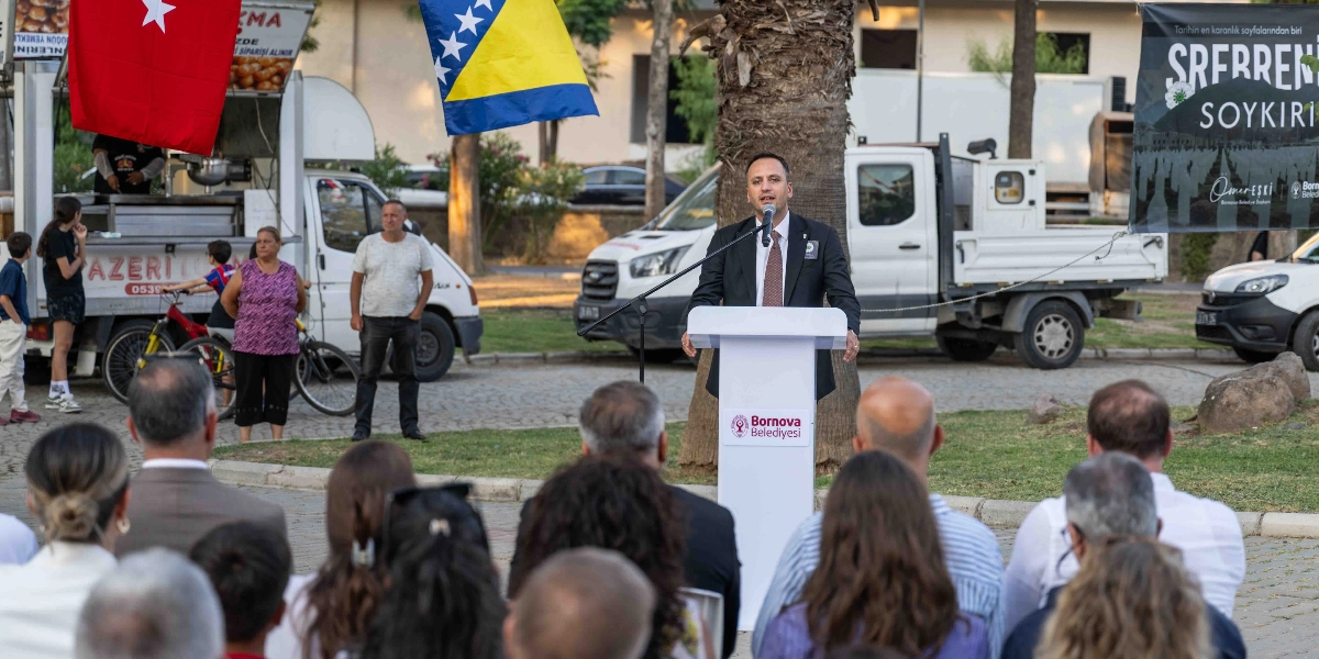 Bornova Belediyesi logolu bir kürsüde konuşma yapan takım elbiseli bir erkek, açık havada düzenlenen anma etkinliğinde dinleyicilere hitap ediyor. Kürsünün arkasında Türkiye ve Bosna Hersek bayrakları asılıyken, sağ tarafta üzerinde 
