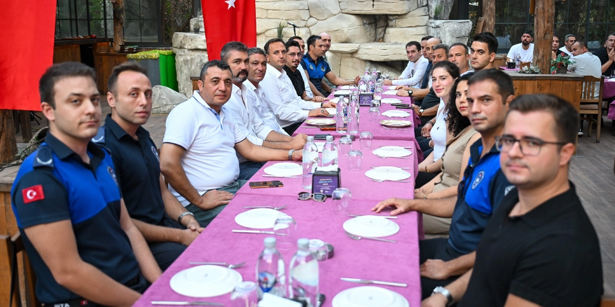 Uzun, mor örtülü bir masanın etrafında toplanmış, aralarında zabıta üniformalı görevlilerin de bulunduğu kalabalık bir grup insan, akşam yemeği için otururken objektife bakıyor. Masanın iki tarafına dizilmiş katılımcıların önünde boş tabaklar, su şişeleri ve çatal bıçak takımları yer alıyor. Arka planda Türk bayrakları ve taş duvarlı bir mekan dekoru görülüyor.