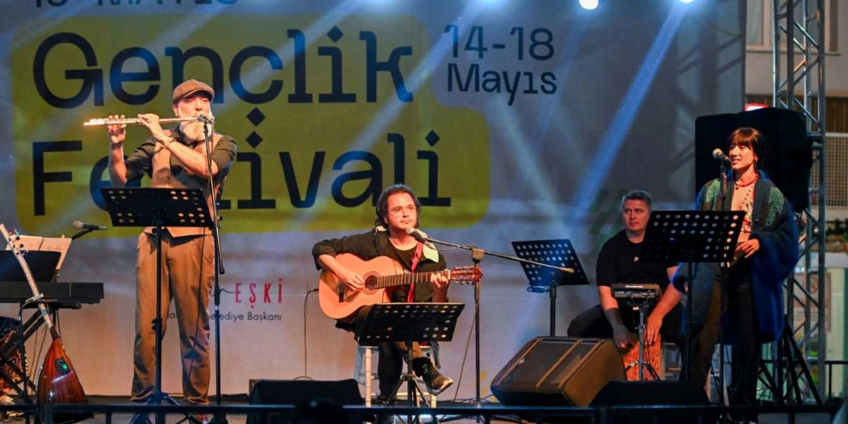 Küçükpark'ta düzenlenen Gençlik Festivali sahnesinde dört kişilik bir müzik grubu performans sergiliyor. Solda ayakta yan flüt çalan sakallı ve kasketli bir müzisyen, ortada oturarak gitar çalan gözlüklü bir sanatçı, sağ tarafta ise elinde mikrofonla şarkı söyleyen bir kadın yer alıyor. Grubun arkasında oturan bir diğer müzisyen ise perküsyon aleti çalıyor. Arka planda üzerinde 
