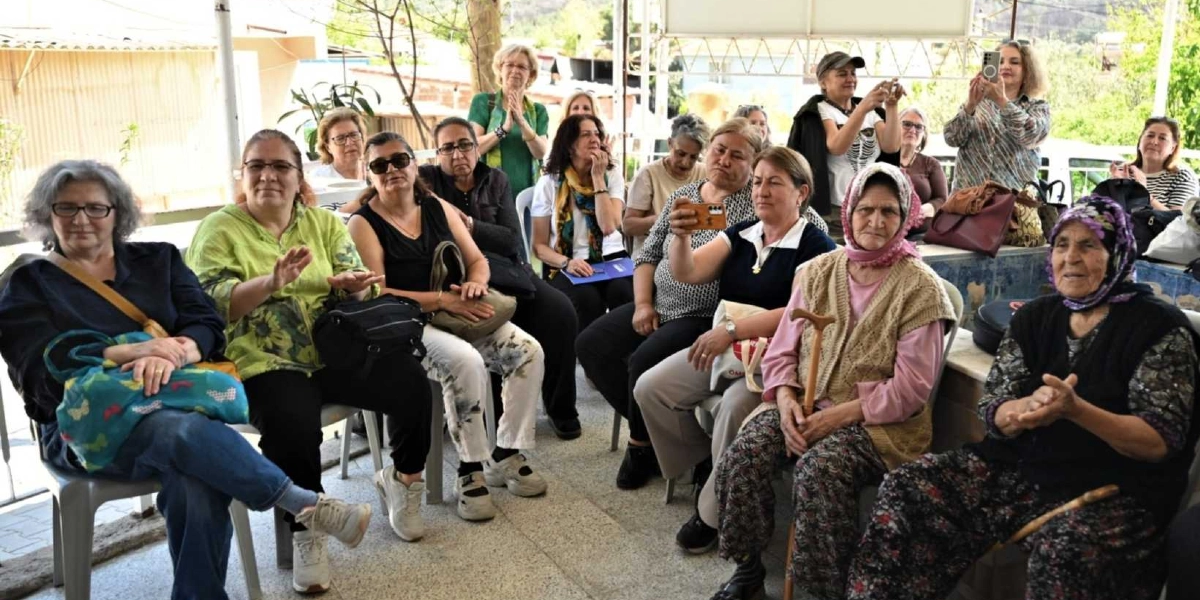 Açık havada, sandalyelerde yan yana oturmuş ve bir etkinliği izleyen, çoğunluğu orta yaş ve üzeri kadınlardan oluşan bir grup. Kadınların bir kısmı alkışlıyor, bazıları ise ellerindeki telefonlarla fotoğraf veya video çekiyor. Ön planda, elinde bastonuyla oturan ve başörtüsü takan yaşlı bir kadın ile yanında yine bastonlu, koyu renkli kıyafetli bir başka kadın dikkat çekiyor. Arka planda ise ayakta duran ve etkinliği takip eden diğer katılımcılar yer alıyor.