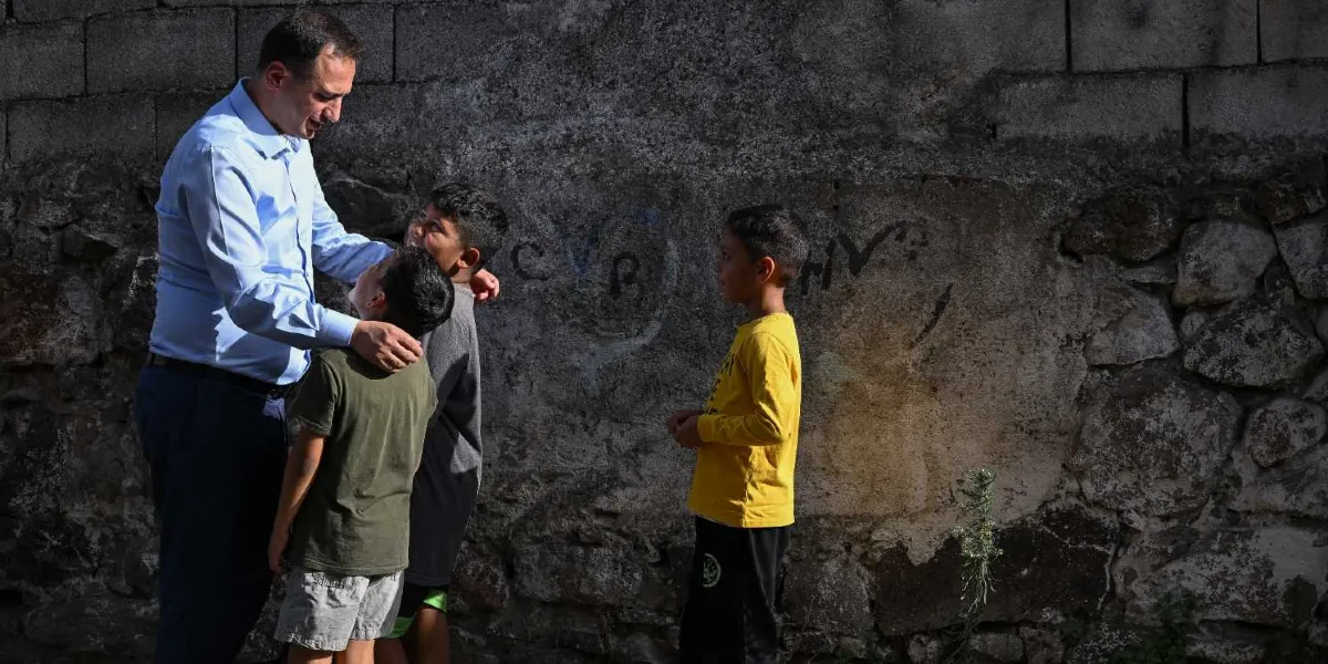 Açık mavi gömlekli bir erkek, taş bir duvarın önünde duran üç küçük çocukla samimi bir şekilde sohbet ediyor. Erkek, çocuklardan birinin omzuna elini koymuş, çocuklar ise ona sevgiyle bakıyor. Çocuklardan biri sarı uzun kollu bir tişört, diğerleri ise kısa kollu tişörtler giyiyor.