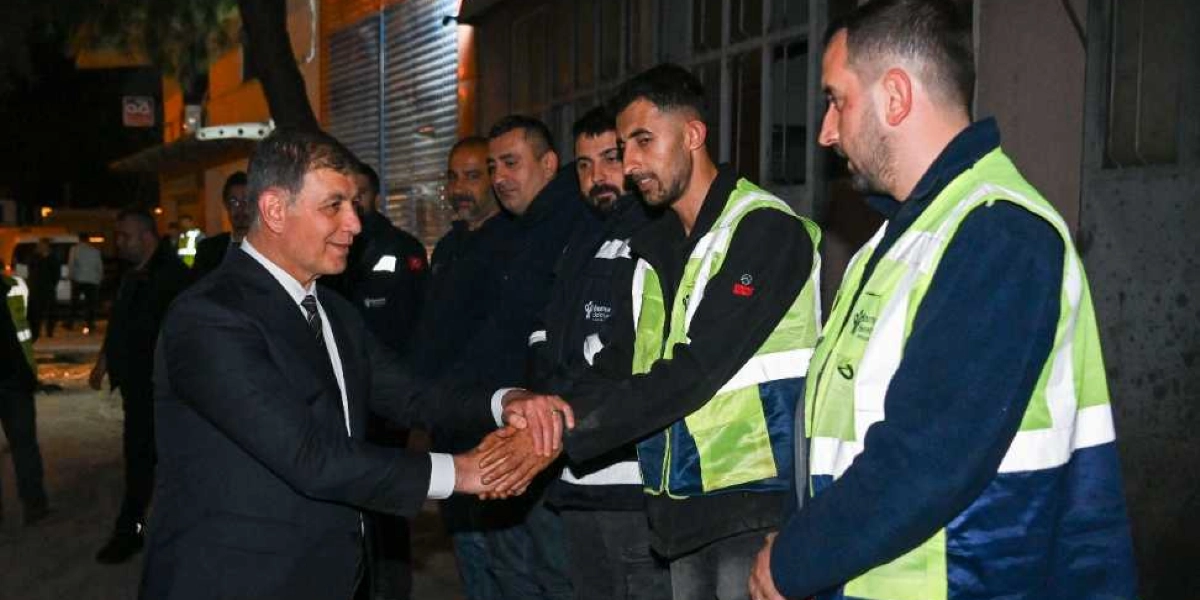 Koyu renk takım elbiseli bir yetkili, gece vakti dışarıda çalışan ve üzerinde reflektörlü iş yelekleri bulunan bir grup işçiyle tokalaşıyor. İşçilerin bir kısmı yeleklerinin üzerinde belediye logosu taşırken, grup samimi bir şekilde bir araya gelmiş durumda.