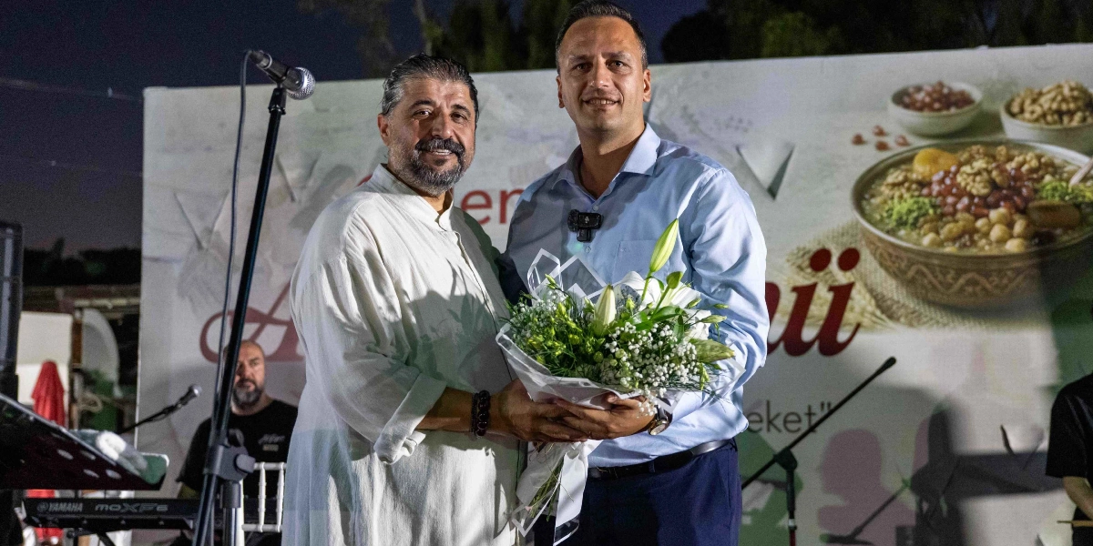 Açık renkli, yakasız bir gömlek giyen sakallı bir erkek, açık mavi gömlekli ve lacivert pantolonlu bir erkeğe beyaz çiçeklerden oluşan bir buket takdim ediyor. İkili, üzerinde aşure kasesi görseli ve 