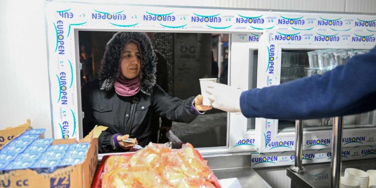 Kışlık montlu ve kapüşonlu bir kadın, beyaz plastik pencereli bir bölmeden dışarıya doğru elinde tuttuğu bir bardak sıcak içeceği uzatıyor. Pencerenin önündeki tezgahta paketlenmiş yiyecekler ve kutu içecekler bulunuyor. Karşı tarafta ise beyaz eldivenli bir kişi, kadının uzattığı bardağı teslim alıyor.