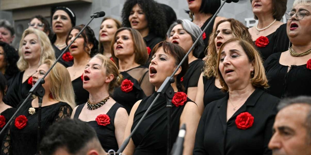 Siyah kıyafetler giymiş ve yakalarında kırmızı gül takılı olan bir koro, Çanakkale Zaferi anma etkinliği kapsamında sahnede şarkı söylüyor. Mikrofonlar önünde duran kadın ağırlıklı grup, ciddiyetle ve coşkuyla performans sergiliyor.