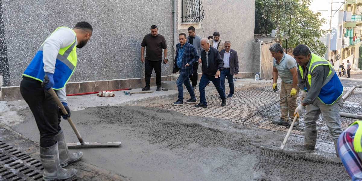 İş yeleği giymiş iki işçi, ellerindeki aletlerle yeni dökülen betonu düzeltirken, arkalarında resmi kıyafetli bir grup erkek çalışmaları inceleyerek yürüyor.