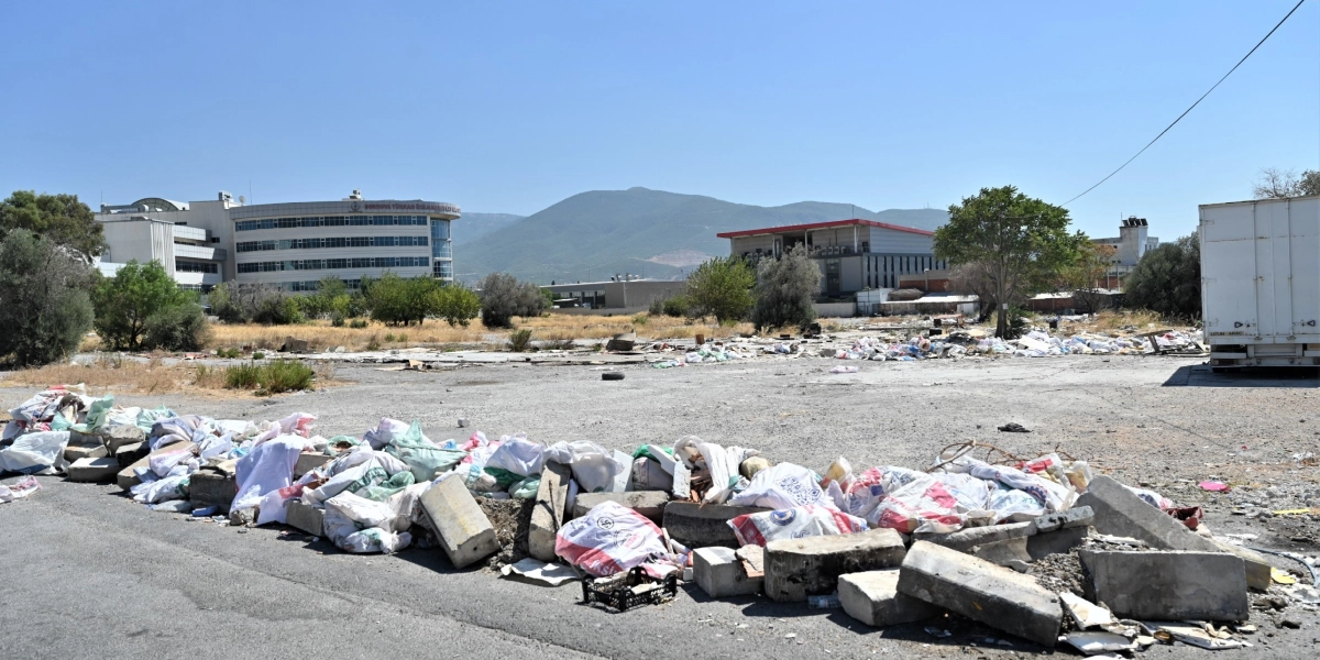 Açık bir arazide, ön planda yol kenarına yığılmış çok sayıda moloz dolu çuval, beton parçaları ve inşaat atıkları bulunuyor. Arka planda Bornova Türkan Özilhan Devlet Hastanesi binası, ağaçlar ve dağ manzarası görülüyor. Sağ tarafta ise beyaz bir tır dorsesi yer alıyor.