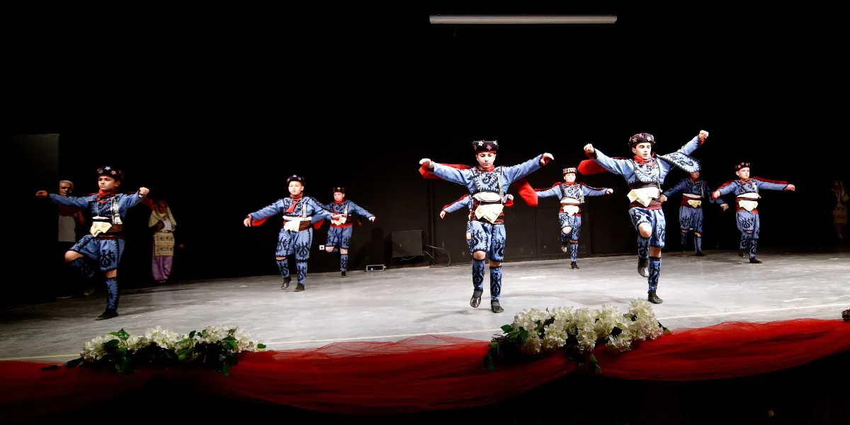 Siyah bir sahne üzerinde, geleneksel mavi işlemeli kıyafetler ve başlıklar giymiş yedi çocuk, halk oyunu sergiliyor. Çocuklar kollarını iki yana açmış, senkronize bir şekilde dans ediyorlar. Sahnenin ön kısmında beyaz çiçekler ve kırmızı tül süslemeler yer alıyor.