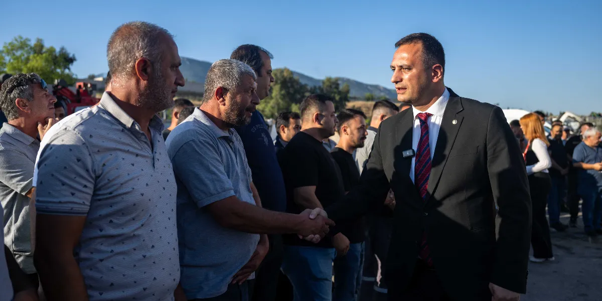 Açık havada, güneşli bir günde siyah takım elbiseli ve kırmızı çizgili kravatlı bir erkek, gri polo yaka tişört giyen bir adamla tokalaşıyor. Çevredeki diğer erkekler bu anı izlerken, arka planda ağaçlıklı bir tepe ve açık bir gökyüzü görülüyor.