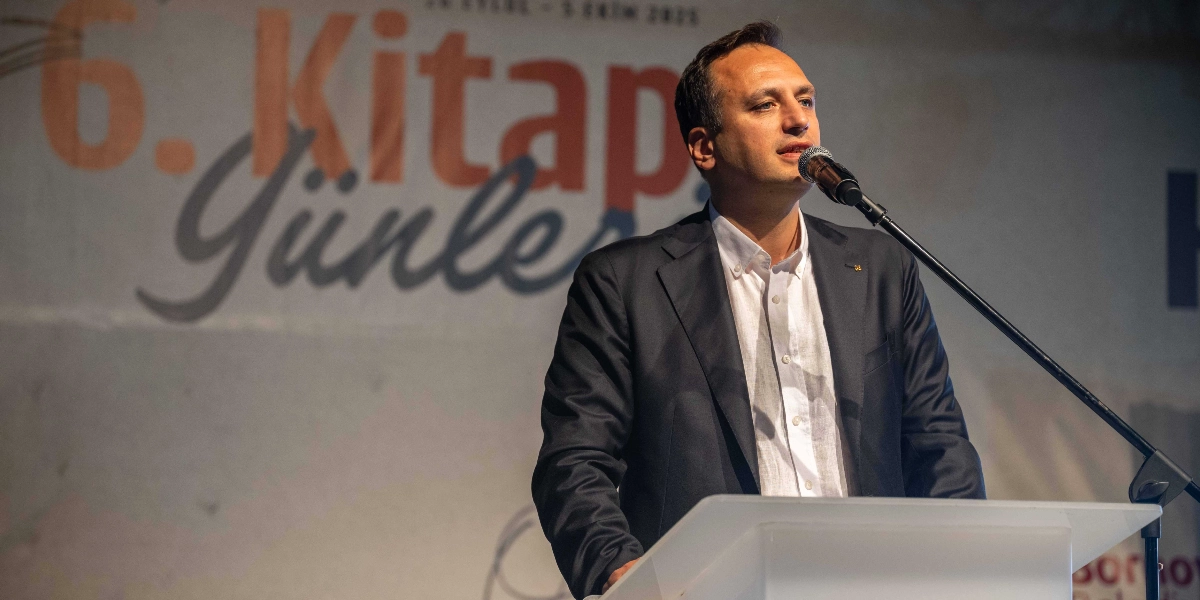 Koyu renk ceket ve beyaz gömlek giymiş bir erkek, kürsüde mikrofon başında konuşma yapıyor. Arka planda üzerinde 