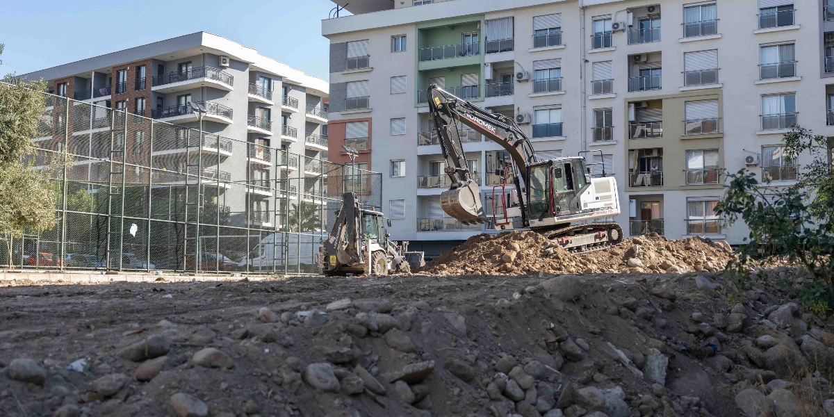 Bornova Erzene'de çok katlı apartmanların önündeki boş arazide, bir paletli ekskavatör ve bir kepçeli iş makinesi toprak kazı çalışması yapıyor.