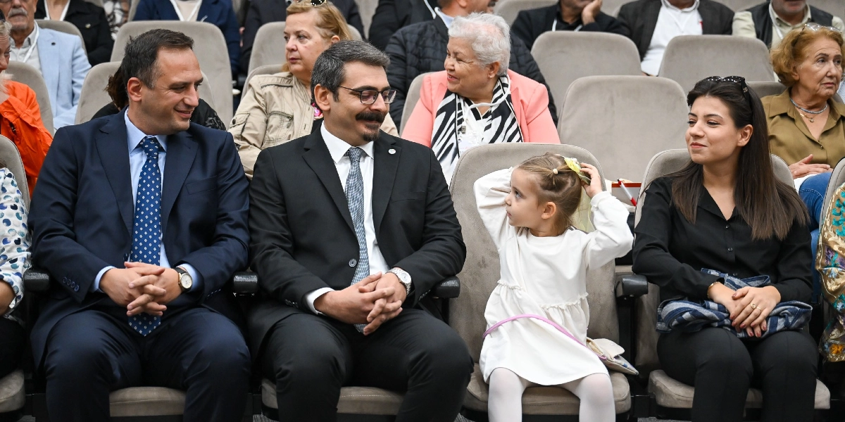 Konferans salonunda yan yana oturan bir grup insan; ön sırada takım elbiseli iki erkek, yanlarında beyaz elbiseli küçük bir kız çocuğu ve siyah kıyafetli bir kadın bulunuyor. Küçük kız çocuğu elleriyle saçındaki tokayı düzeltirken, yanındaki yetişkinler gülümseyerek ona bakıyor. Arka sıralarda ise çeşitli yaş gruplarından izleyiciler oturuyor.