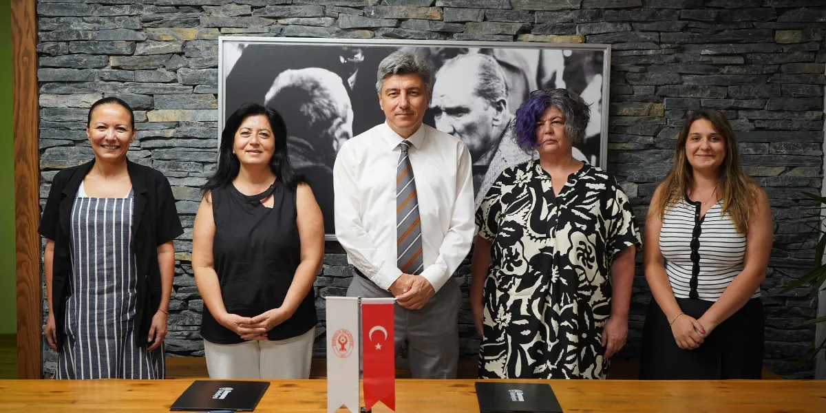 Taş duvarlı bir arka plan önünde, Mustafa Kemal Atatürk'ün siyah beyaz bir portresinin altında yan yana duran dört kadın ve bir erkek. Ortada beyaz gömlekli ve çizgili kravatlı bir erkek, yanında ise günlük kıyafetler içindeki dört kadın yer alıyor. Önlerindeki ahşap masanın üzerinde Bornova Belediyesi logosunun bulunduğu küçük bir flama ve Türk bayrağı ile imzalanmayı bekleyen belgeler bulunuyor.