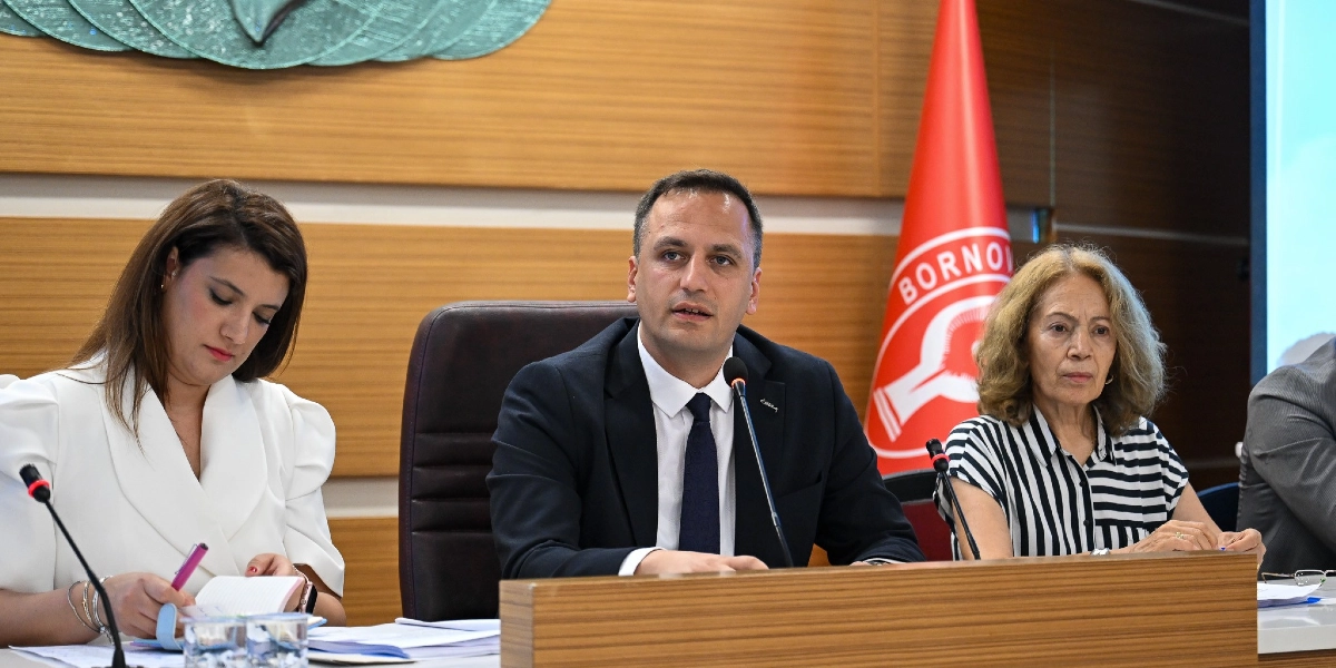 Toplantı masasında oturan üç kişi, önlerinde mikrofonlar ve notlar ile bir sunum yapıyor. Ortada koyu renk takım elbiseli ve kravatlı bir erkek konuşurken, solunda beyaz bluzlu bir kadın not alıyor, sağında ise çizgili bluzlu bir kadın oturuyor. Arka planda üzerinde Bornova Belediyesi logosu bulunan kırmızı bir bayrak ve ahşap panelli bir duvar yer alıyor.