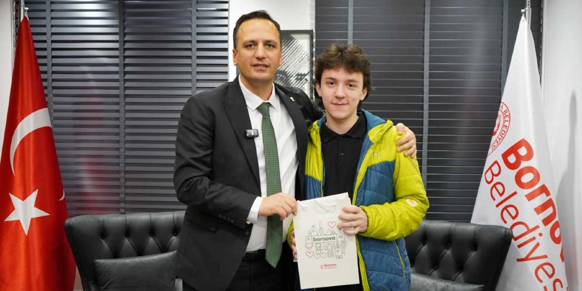 Bornova Belediye Başkanı, yanında duran genç bir öğrenciye üzerinde Bornova Belediyesi logosu ve çizimleri bulunan beyaz bir hediye çantası veriyor. İkili, arkalarında Türk bayrağı ve Bornova Belediyesi bayrağı bulunan bir ofis ortamında yan yana durarak gülümsüyor.