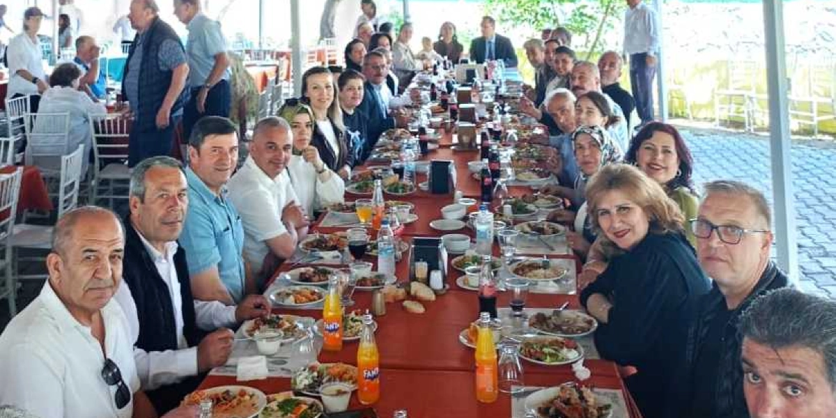 Açık havada, uzun bir yemek masasında bir araya gelmiş kalabalık bir grup insan, önlerindeki tabaklarda yemek yiyor ve sohbet ediyor. Masanın üzerinde içecekler, çeşitli yemek tabakları ve servisler bulunuyor. Katılımcılar arasında muhtarların ve yerel temsilcilerin yer aldığı, samimi bir ortamda gerçekleşen bir etkinlik görüntüsü.