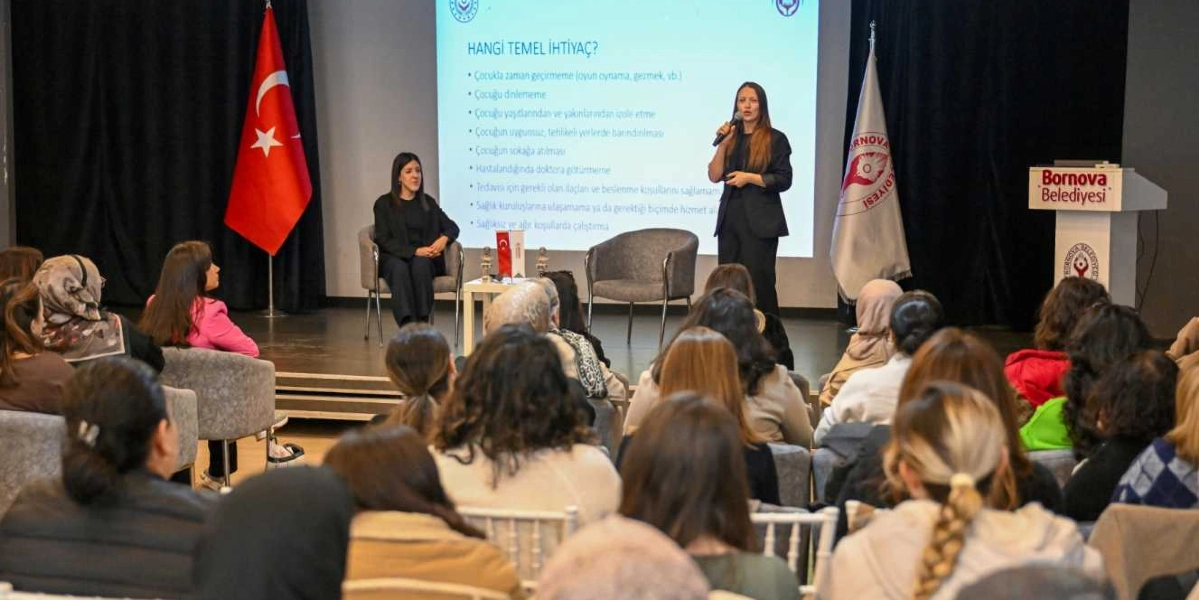 Bir konferans salonunda, sahnedeki bir kadın konuşmacı mikrofonla sunum yaparken, yanında oturan bir diğer kadın onu dinliyor. Arkalarındaki projeksiyon ekranında 
