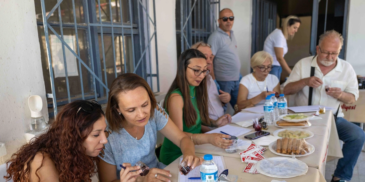 Bornova Reçel Şenliği'nde jüri üyeleri, önlerindeki masada bulunan küçük kavanozlardaki reçelleri tadarak puanlama yapıyor. Masada çeşitli reçel örnekleri, su şişeleri, not kağıtları ve üzerinde 