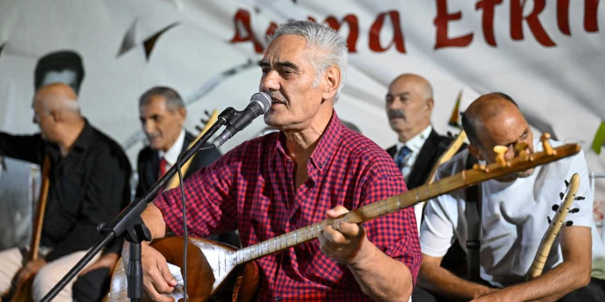 Kırmızı ekose gömlekli, gri saçlı bir halk ozanı, elinde bağlamasıyla mikrofon başında şarkı söylüyor. Arka planda ona eşlik eden, bağlama çalan diğer müzisyenler yer alıyor. Arkadaki beyaz pankart üzerinde kısmen görünen 