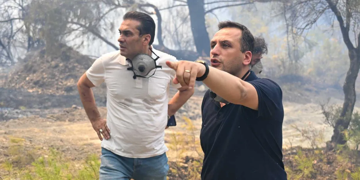 Yangın sonrası küle dönmüş bir arazide, boyunlarında koruyucu maskelerle duran iki erkekten biri, eliyle uzak bir noktayı işaret ederek bir şeyler anlatıyor. Arka planda dumanların yükseldiği, ağaçların yandığı ve toprağın karardığı bir ormanlık alan görülüyor.