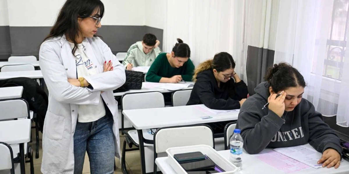 Sınıf ortamında deneme sınavına giren öğrenciler ve onları gözlemleyen bir öğretmen. Öğrenciler sıralarında oturmuş, önlerindeki sınav kağıtlarına odaklanmış durumdalar. Beyaz önlüklü bir kadın öğretmen, kollarını kavuşturmuş bir şekilde öğrencilerin çalışmalarını takip ediyor. Masaların üzerinde sınav kağıtları, su şişeleri ve kişisel eşyalar bulunuyor.