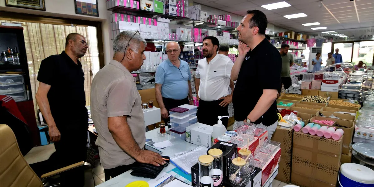 Bej renkli gömlek giyen bir adamın, siyah tişörtlü bir erkekle ve yanındaki diğer kişilerle bir dükkanın içinde sohbet ettiği an. Dükkanın raflarında ve masanın üzerinde çeşitli cam mutfak eşyaları, saklama kapları ve hediyelik ürünler sergileniyor. Arka planda dükkan çalışanları ve diğer müşteriler görülüyor.