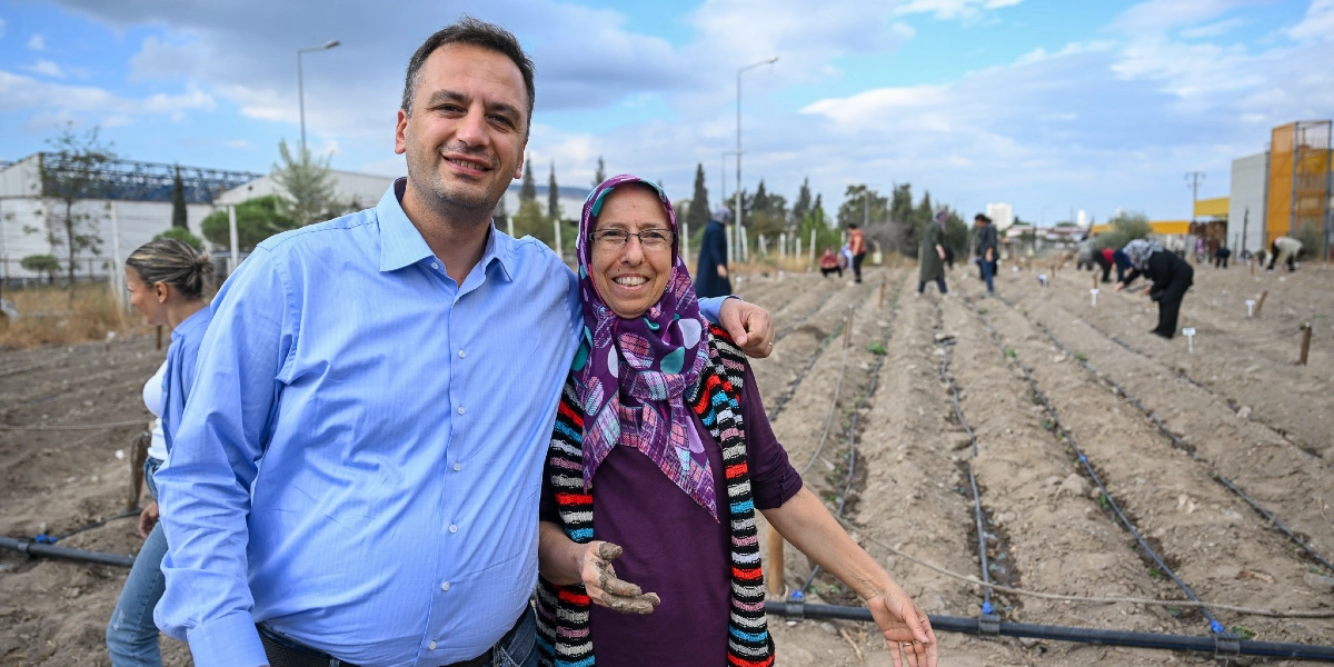 Açık havada, ekili bir tarım arazisinde yan yana duran, gülümseyen bir erkek ve başörtülü bir kadın. Kadın, elleri toprakla kirlenmiş bir şekilde, üzerinde mor bir bluz ve renkli çizgili bir hırka ile poz veriyor. Arka planda ise toprağı işleyen diğer kadınlar ve damlama sulama boruları görülüyor.