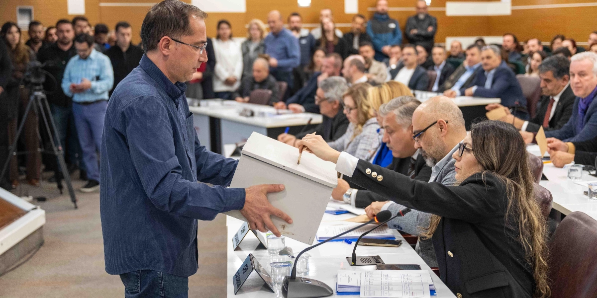 Bornova Belediye Meclisi toplantısında, lacivert gömlekli bir erkek elinde tuttuğu beyaz oy sandığına, uzun saçlı ve siyah ceketli bir kadın üyenin oy pusulasını atmasını bekliyor. Toplantı salonunda masaların etrafında oturan diğer meclis üyeleri ve ayakta izleyen katılımcılar yer alıyor.