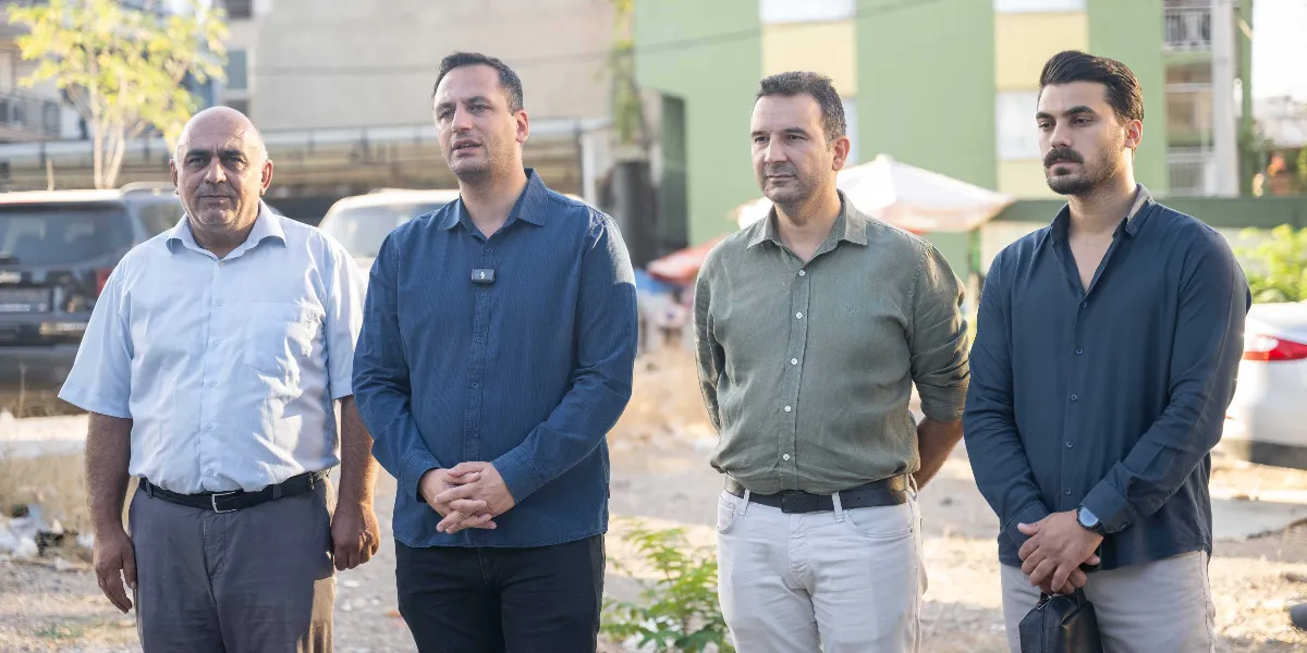 Açık havada, yan yana duran dört erkek; soldan sağa doğru açık mavi kısa kollu gömlekli, lacivert uzun kollu gömlekli, haki renkli uzun kollu gömlekli ve koyu lacivert uzun kollu gömlekli kişiler, ciddi bir ifadeyle karşıya bakıyor.