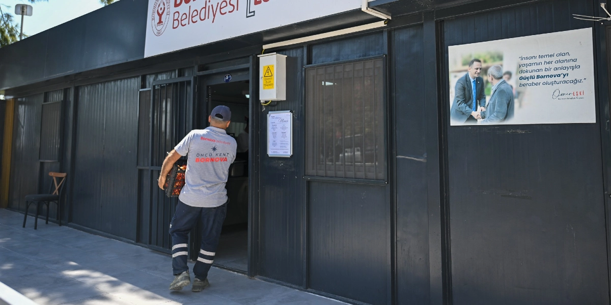 Bornova Belediyesi logolu gri tişörtlü bir çalışan, elinde bir kasa dolusu ürünle siyah renkli prefabrik bir binanın girişinden içeri giriyor. Binanın dış cephesinde, üzerinde Bornova Belediye Başkanı Ömer Eşki'nin bir vatandaşla tokalaştığı fotoğrafın ve 