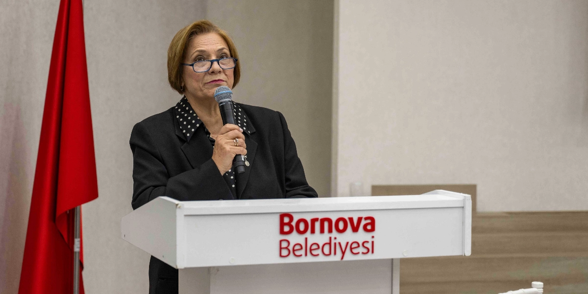 Bornova Belediyesi yazılı kürsüde konuşma yapan, gözlüklü, siyah ceketli ve puantiyeli gömlek giymiş orta yaşlı kadın. Arka planda kırmızı bir Türk bayrağı yer alıyor.