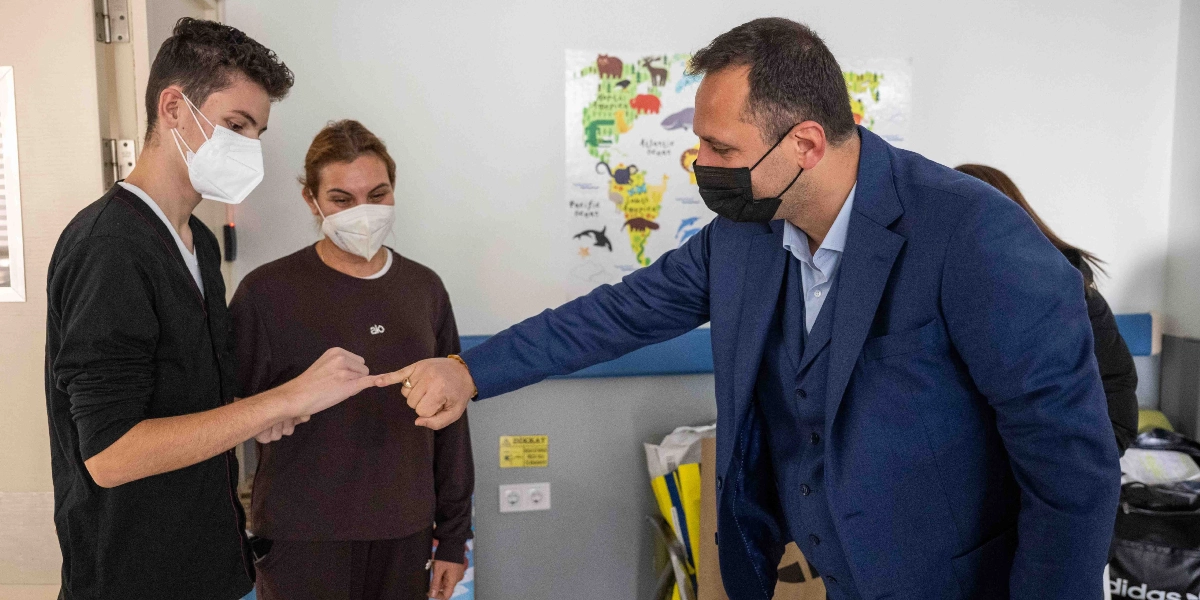 Lacivert takım elbiseli ve siyah maskeli bir erkek, karşısında duran genç bir erkekle serçe parmaklarını birbirine kenetleyerek sözleşme yapıyor. Genç erkeğin yanında, üzerinde 