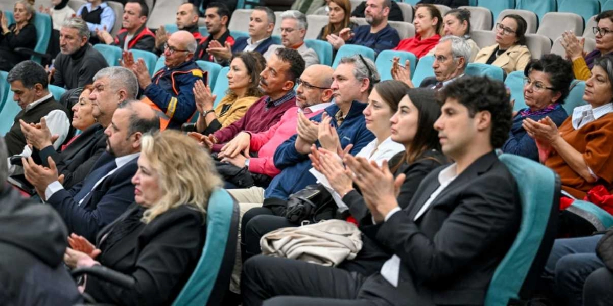 Konferans salonunda oturan ve alkışlayan kalabalık bir izleyici grubu. Farklı yaş ve meslek gruplarından oluşan katılımcılar, turkuaz renkli koltuklarda yan yana oturarak sahnedeki bir konuşmayı veya etkinliği dikkatle takip ediyor. Ön planda takım elbiseli erkekler ve çeşitli kıyafetler içindeki kadınlar yer alırken, arka sıralarda daha fazla katılımcı görülüyor. Ortam resmi bir toplantı havasını yansıtıyor.