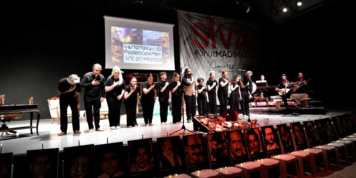Siyah giyinmiş bir grup sanatçı, sahnede ellerini kalplerine koyarak saygı duruşunda bulunuyor. Sahnenin arka planında üzerinde 