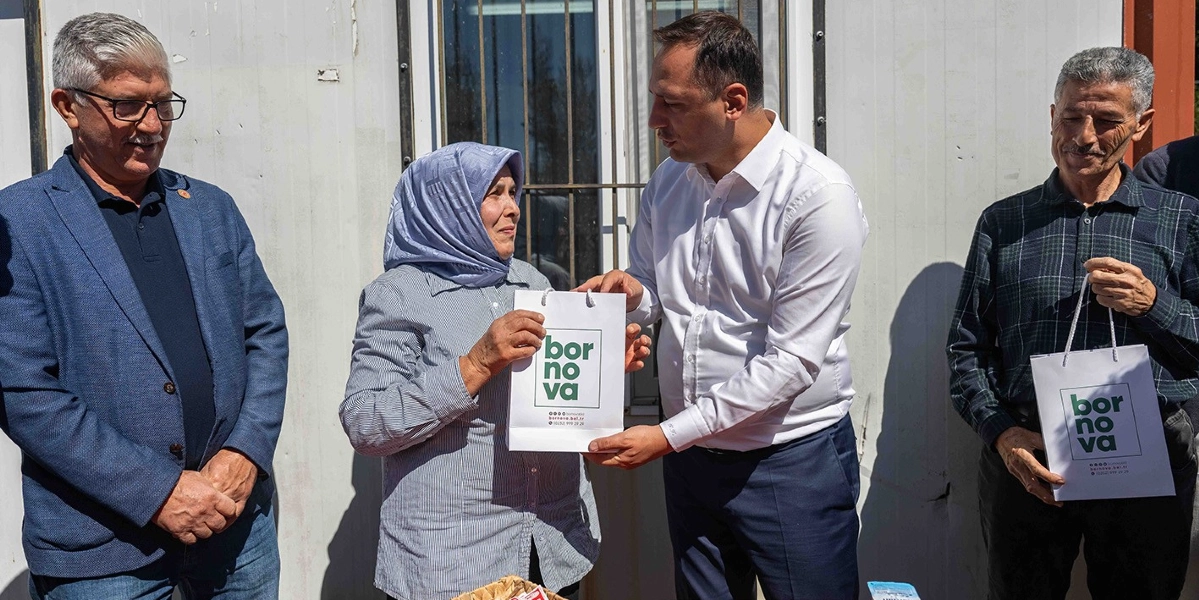 Beyaz gömlekli bir yetkili, üzerinde Bornova Belediyesi logosu bulunan hediye çantasını, başörtülü bir kadına takdim ediyor. Yanlarında iki erkek daha bulunuyor ve hepsi gülümsüyor. Sağ taraftaki kişinin elinde de aynı logolu hediye çantasından bulunuyor. Arka planda beyaz bir duvar ve pencere yer alıyor.