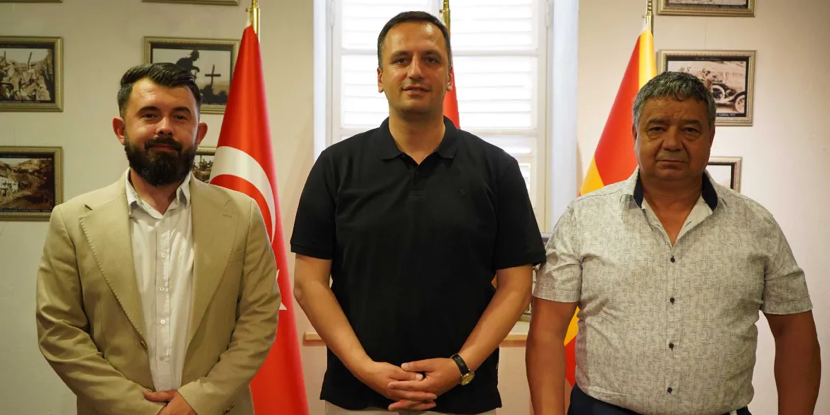 İç mekanda, arkalarında Türkiye ve Kuzey Makedonya bayraklarının bulunduğu, duvarda çerçeveli fotoğrafların yer aldığı bir ortamda yan yana duran üç erkek. Solda bej renkli ceket ve beyaz gömlek giymiş sakallı bir adam, ortada siyah polo yaka tişörtlü bir adam ve sağda kısa kollu desenli bir gömlek giymiş orta yaşlı bir adam yer alıyor. Üçü de kameraya bakarak poz veriyor.